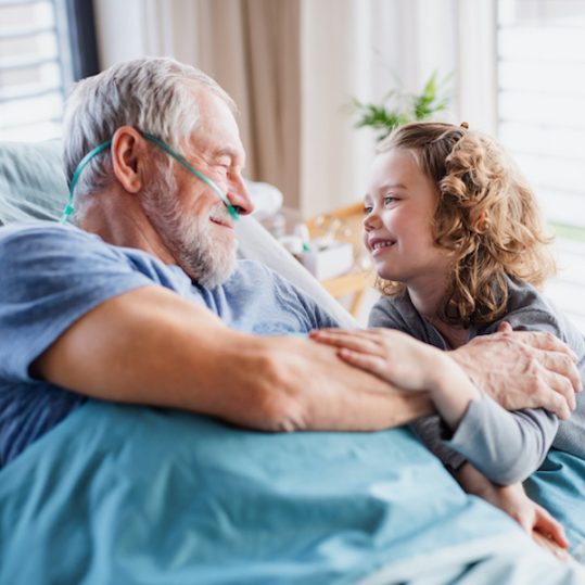A cheerful small girl visiting ill grandfather in bed in hospital room.